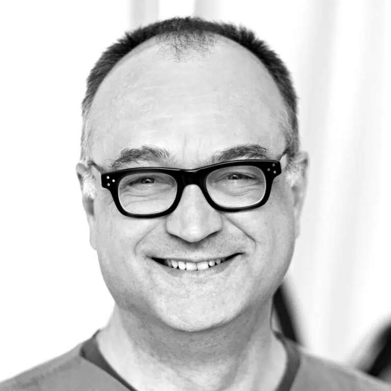 Ein lächelnder Mann mit Brille und kurzem, dunklem Haar vor hellem Hintergrund