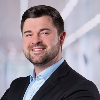 Ein junger Mann mit dunklem Haar und Bart lächelt freundlich im Anzug vor unscharfem Hintergrund