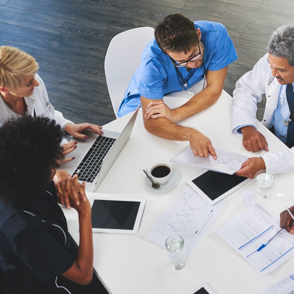Ein Team von Medizinern sitzt mit Laptops um einen Tisch herum. Ärzte und Mitarbeiter besprechen Unterlagen und Testergebnisse. Experten des Gesundheitswesens bei der Bewältigung der täglichen Aufgaben und Pflichten