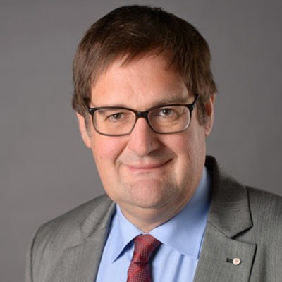 Ein lächelnder Mann mit braunen Haaren und Brille trägt grauen Anzug und rote Krawatte