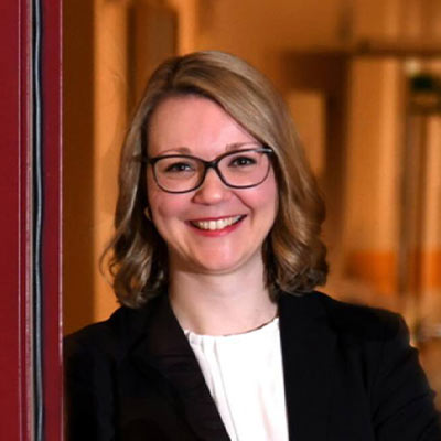 Eine lächelnde Frau mit blonden Haaren und Brille in eleganter dunkler Kleidung