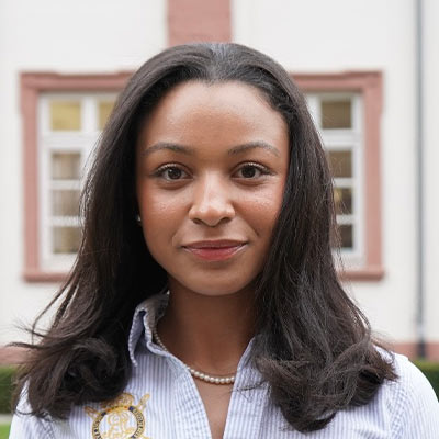 Junge Frau mit dunklen Haaren und Perlenkette lächelt dezent vor hellem Gebäude