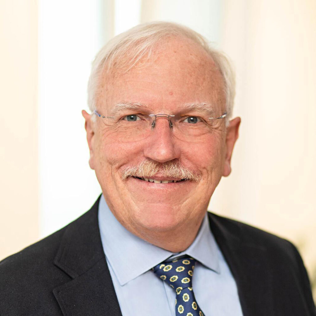 Ein älterer Herr mit grauem Haar und Schnurrbart lächelt freundlich in dunklem Anzug